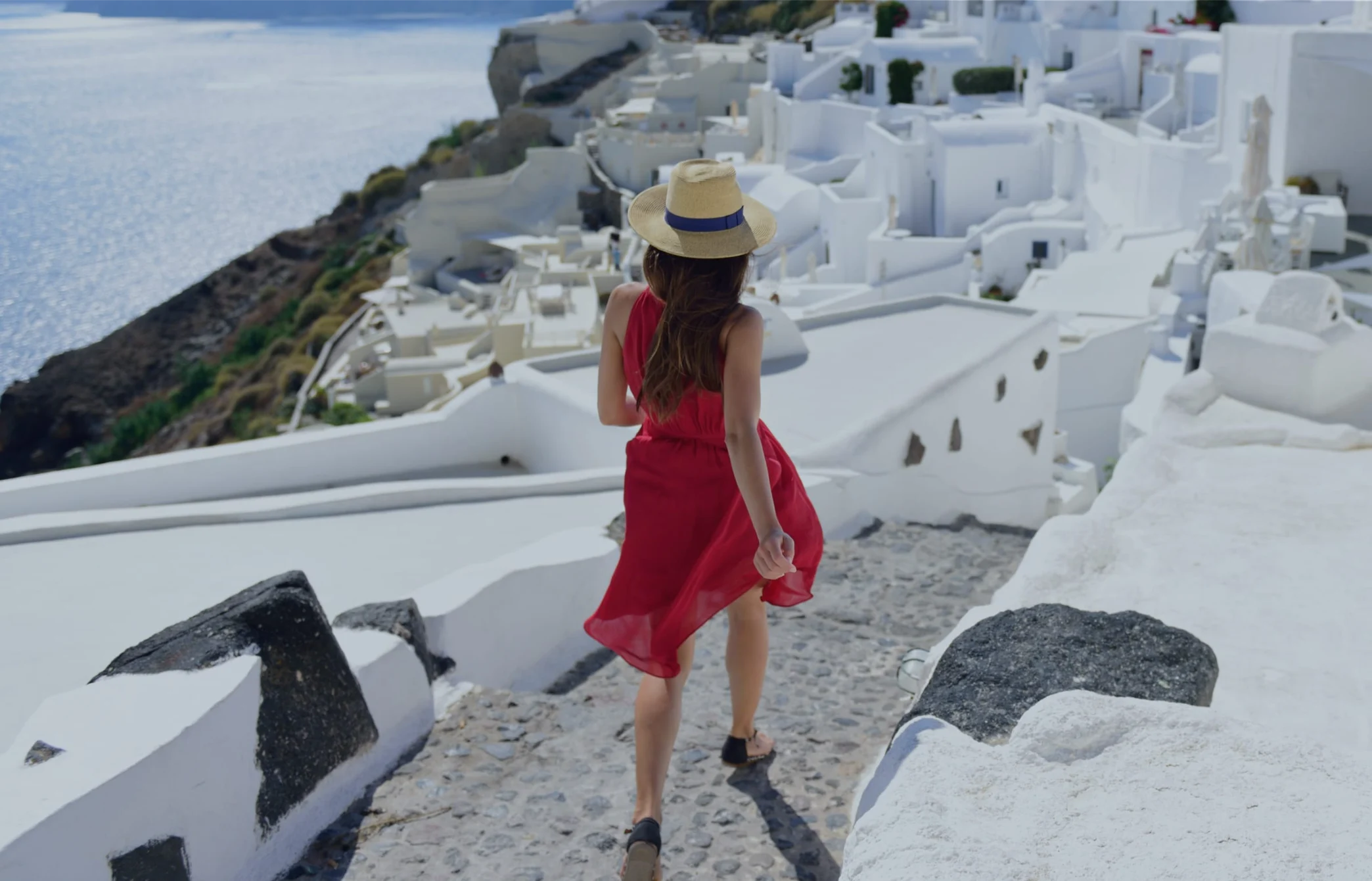 Woman in red dress walking up stone pathway toward whitewashed Greek village buildings overlooking blue sea